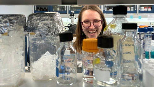charlotte francoeur smiling on the other side of a shelf of glass vials in the lab