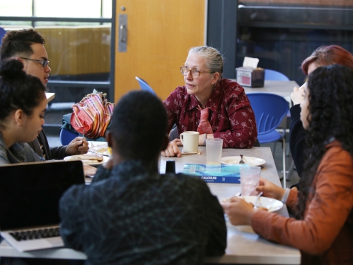 seated professor lecturing at a table where several students are also seated